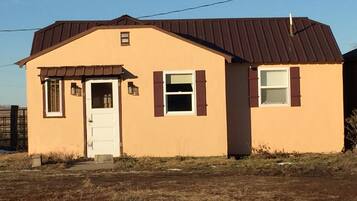 A single-story house with a brown roof and beige walls, featuring a front door, two windows with shutters, and a small porch area.