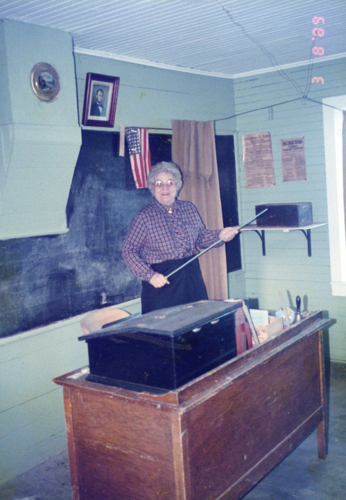 A room with a wooden desk, a blackboard, an American flag, and a framed picture on the wall.