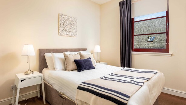 Quarto bem arrumado com cama, duas mesas de cabeceira, uma obra de arte na parede, janela com cortinas e vista para o exterior.