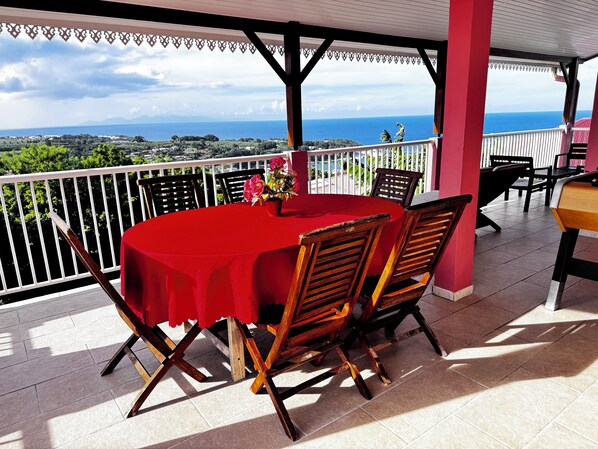 Un espace extérieur abrité, aménagé pour les repas, avec une table et des chaises rouges, offrant une vue panoramique sur l’océan et le paysage.