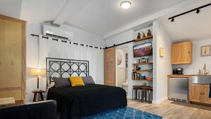 A modern bedroom with a bed, a blue patterned rug, a black sofa, a wooden wardrobe, and a kitchenette with a sink and stove.