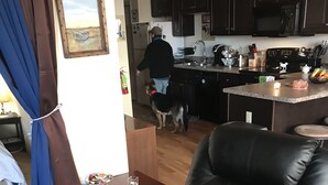 Una sala de estar moderna con un sillón de cuero, una mesita de madera con un control remoto, y una cocina al fondo.
