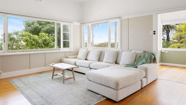 Un salon moderne avec un canapé blanc, une petite table en bois et un tapis gris. De grandes fenêtres offrent une vue sur la verdure à l’extérieur.