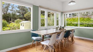 Une salle à manger avec une table et des chaises en bois, de grandes fenêtres donnant sur un paysage verdoyant et un petit abri de jardin à l’extérieur.