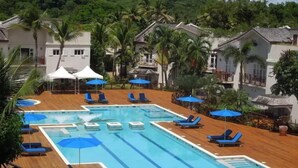A swimming pool with multiple lanes, surrounded by lounge chairs and umbrellas, set against a backdrop of palm trees and buildings. - Cotton Bay Townhouse (Gros Islet)