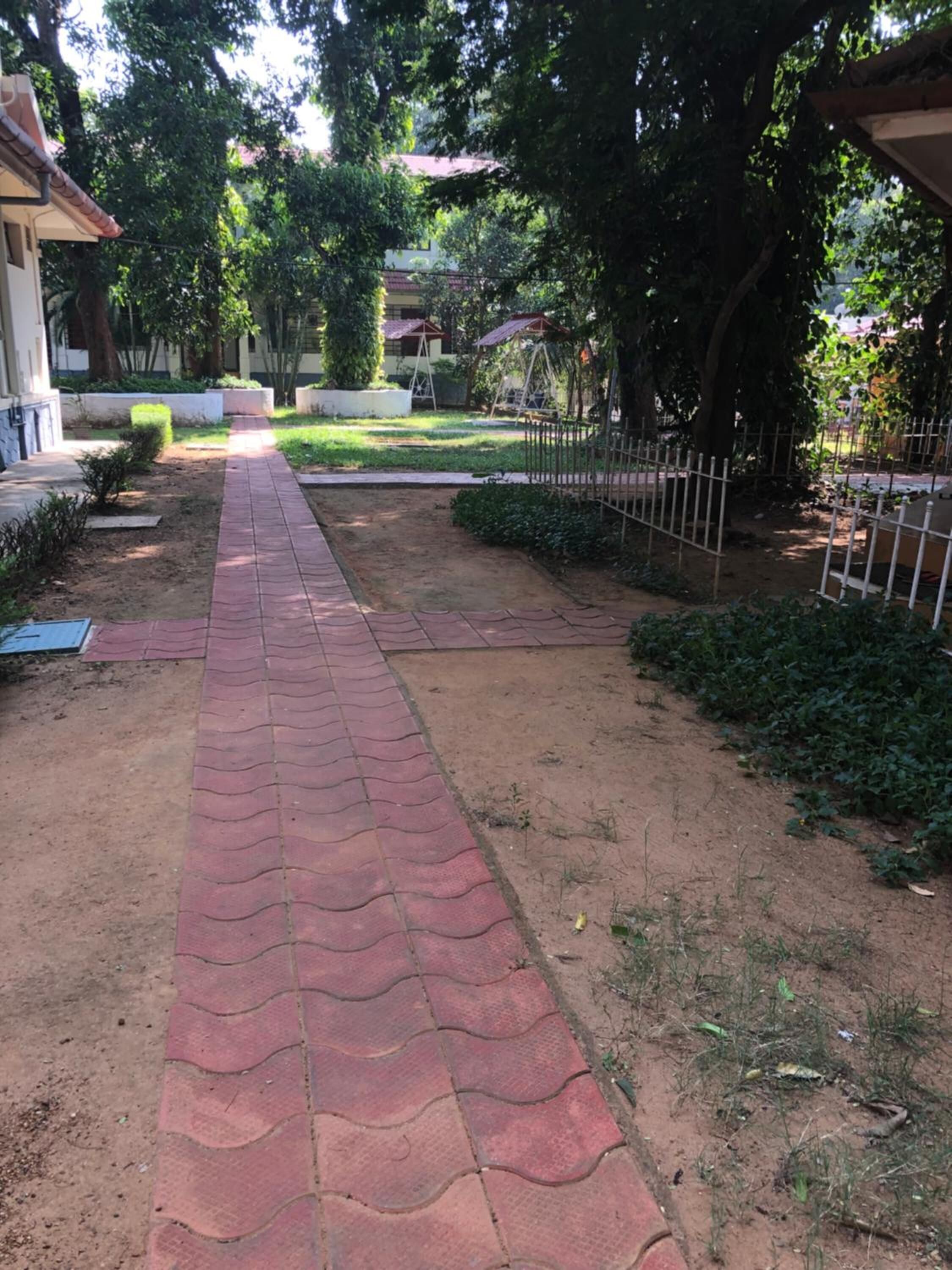 Un chemin pavé traverse un espace aménagé, avec une clôture d’un côté et un bâtiment de l’autre.
