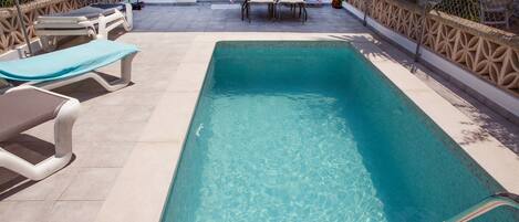 A rectangular swimming pool with clear water, surrounded by lounge chairs and a table with chairs.