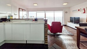 Une cuisine moderne avec un îlot central, une chaise rouge, un téléviseur et une grande fenêtre donnant sur la ville.