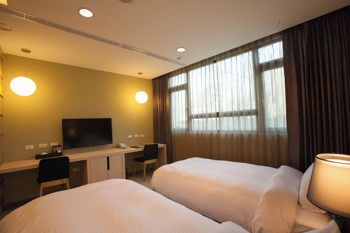 Ein Hotelzimmer mit zwei Betten, einem Schreibtisch mit Stuhl, einem Fernseher und einem Fenster mit Vorhängen.