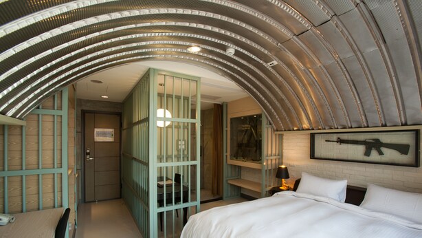 Une chambre d’hôtel dotée d’un plafond incurvé, d’un lit avec une literie blanche, d’un bureau avec une chaise et d’un petit coin salon comprenant une table et des chaises.