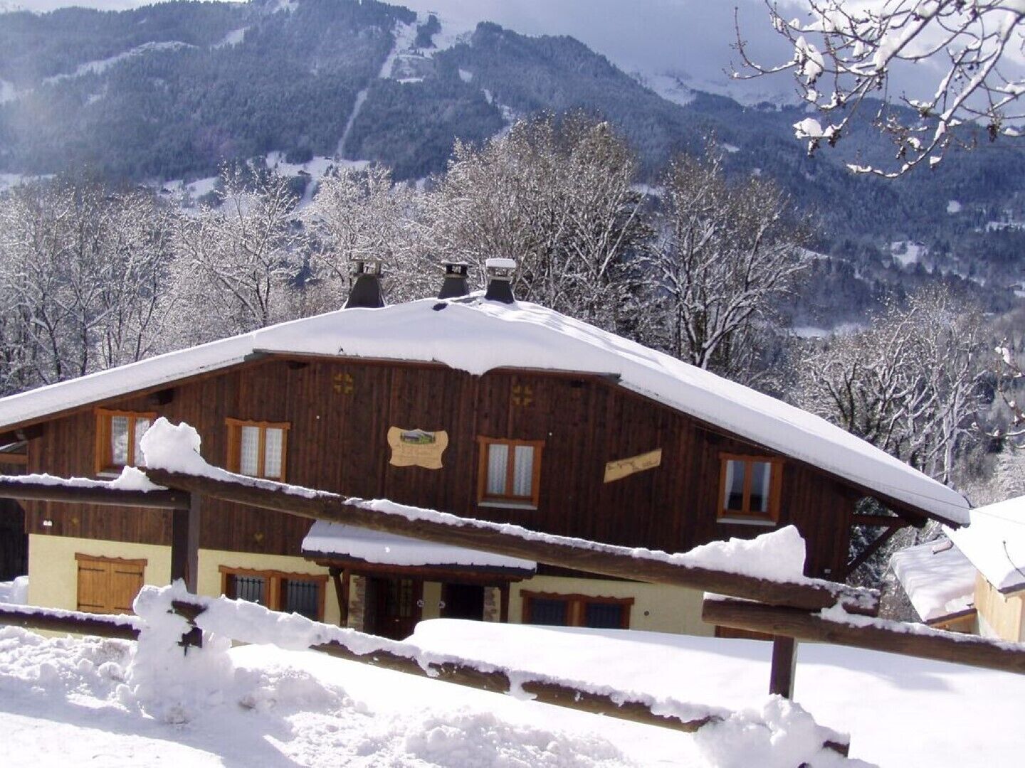 A wooden chalet with a sloped roof covered in snow, surrounded by a snowy landscape and mountains.