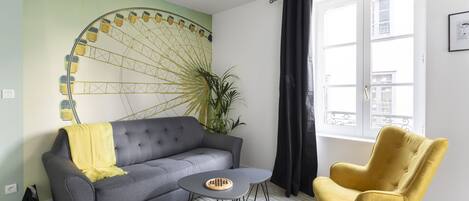 A living room with a grey sofa, a yellow armchair, a small round table, and a large window with black curtains.