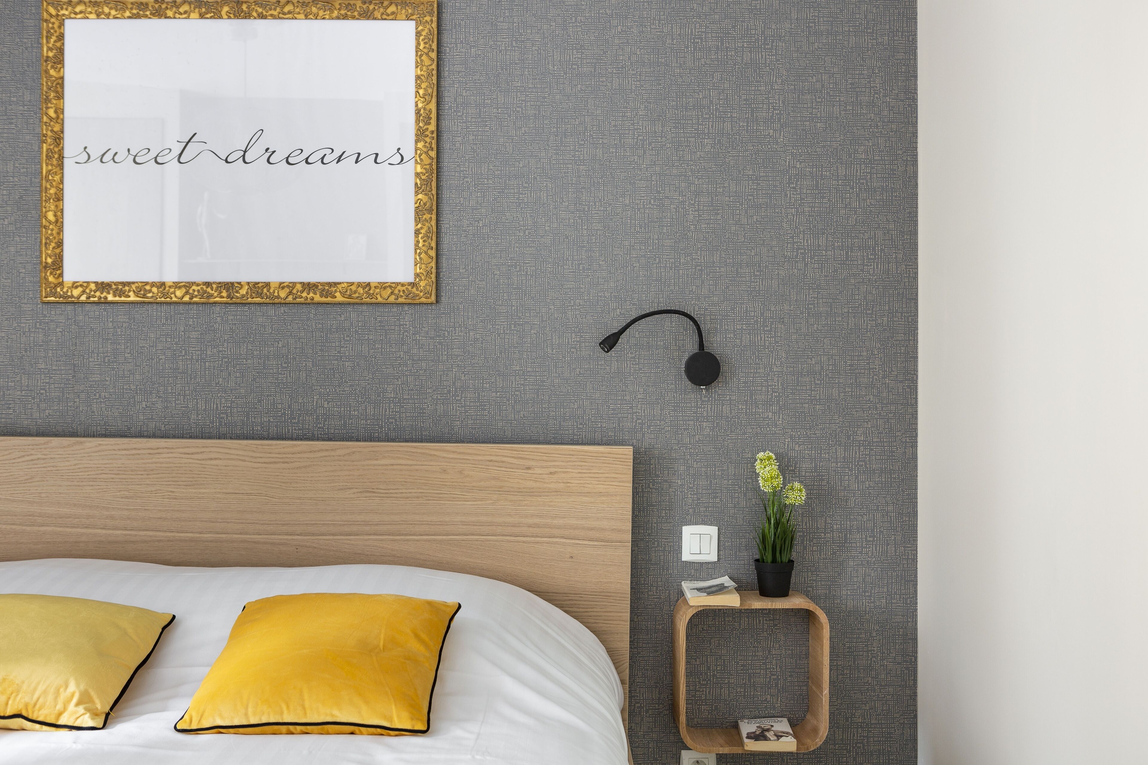 Une chambre à coucher bien rangée, avec un lit, deux oreillers jaunes, une citation inspirante encadrée, une petite table de chevet avec une plante et un haut-parleur fixé au mur.