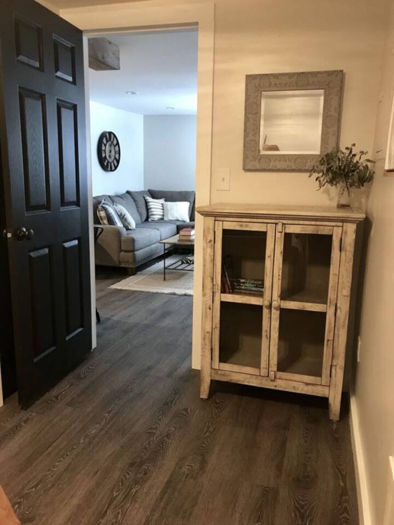 A hallway with a dark wooden floor, a light-colored wall, and a wooden cabinet with glass doors.