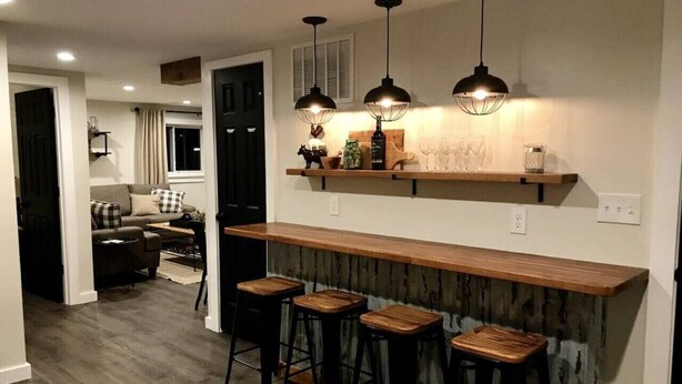 Une cuisine moderne avec un bar en bois, trois tabourets, des luminaires suspendus et un coin repas à l’arrière-plan.