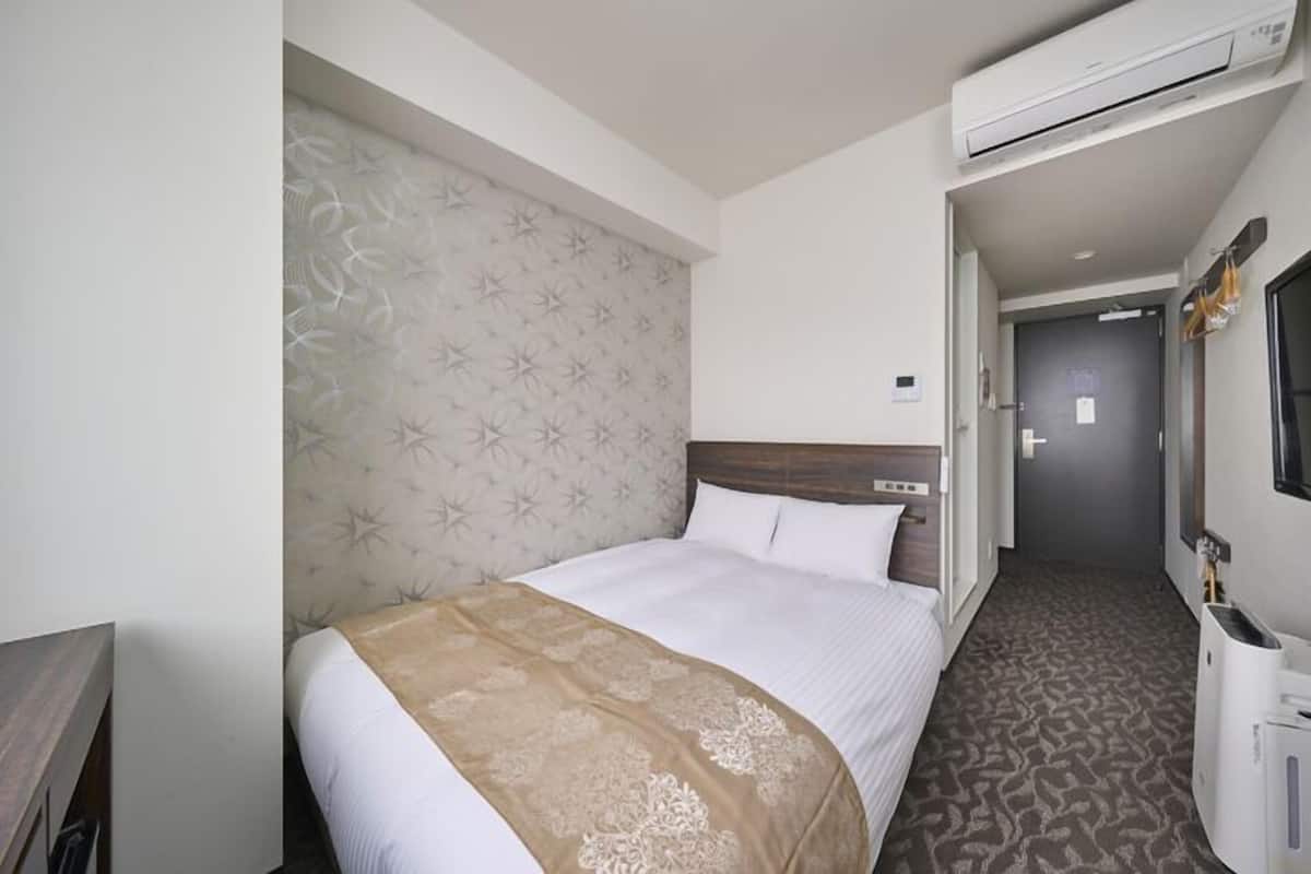 A hotel room with a bed, an air conditioning unit, a bedside table, and a mirror.