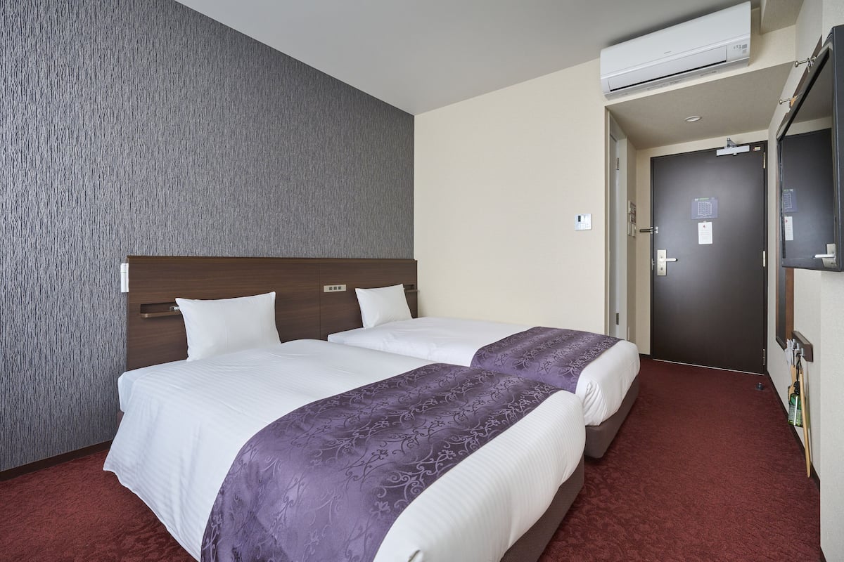 A hotel room with two beds, a grey textured wall, a red carpet, and a door with a mirror and a TV.