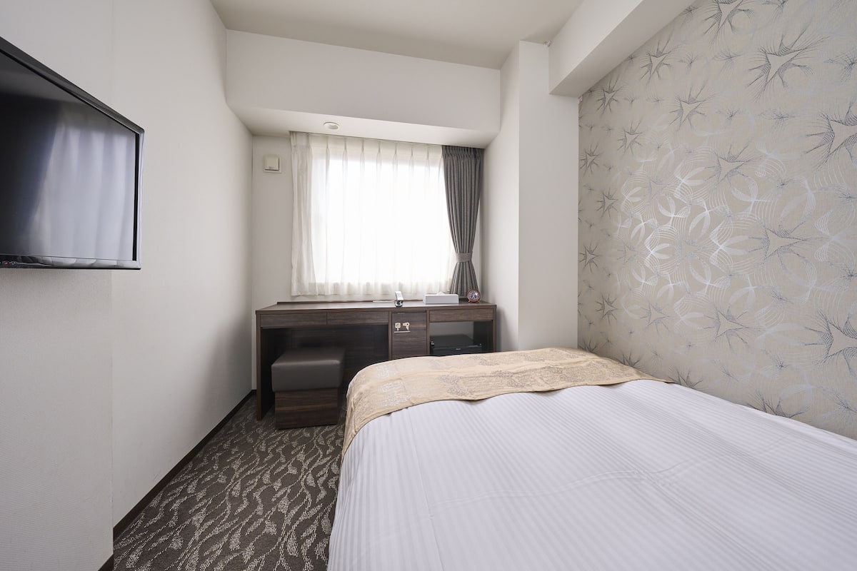 A hotel room with a bed, a desk with a chair, a television, and a window with curtains.