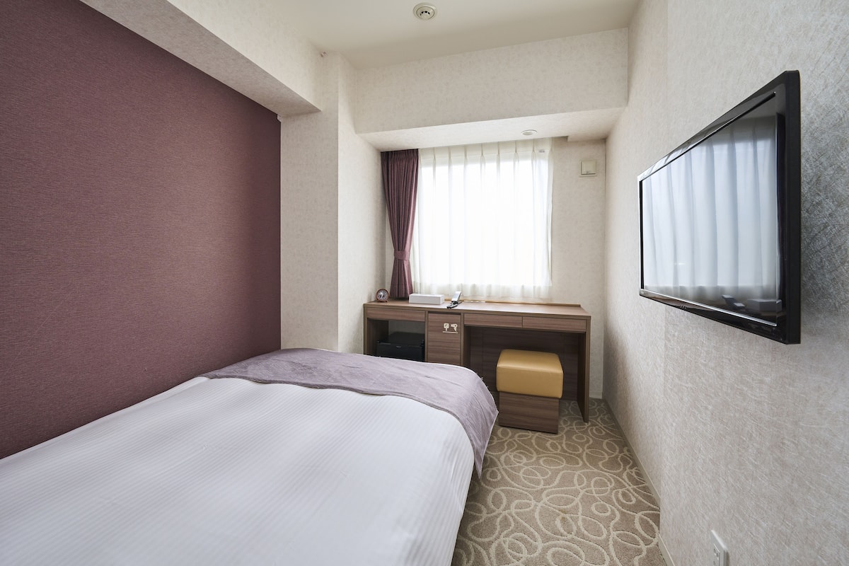 A hotel room with a bed, a desk with a chair, a television, and a window with curtains.
