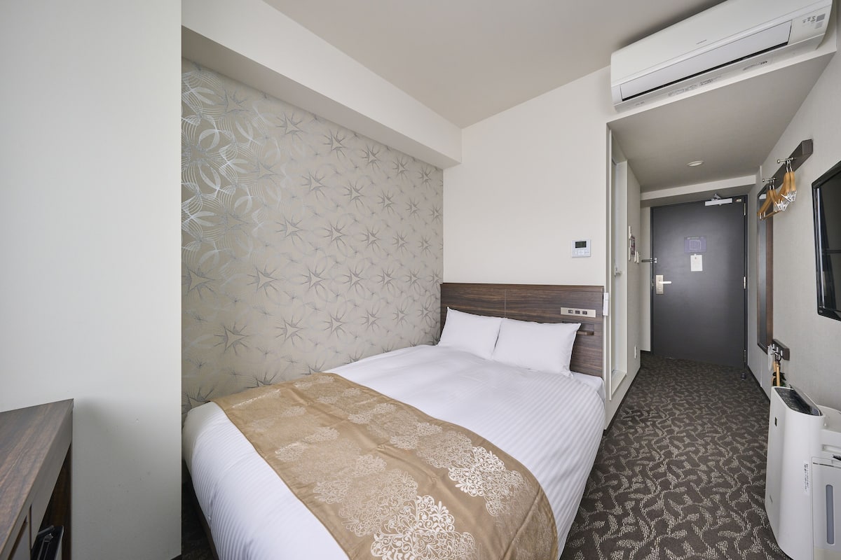 A hotel room with a bed, a headboard with a star pattern, a brown throw blanket, a wooden nightstand, and a wall-mounted air conditioner.