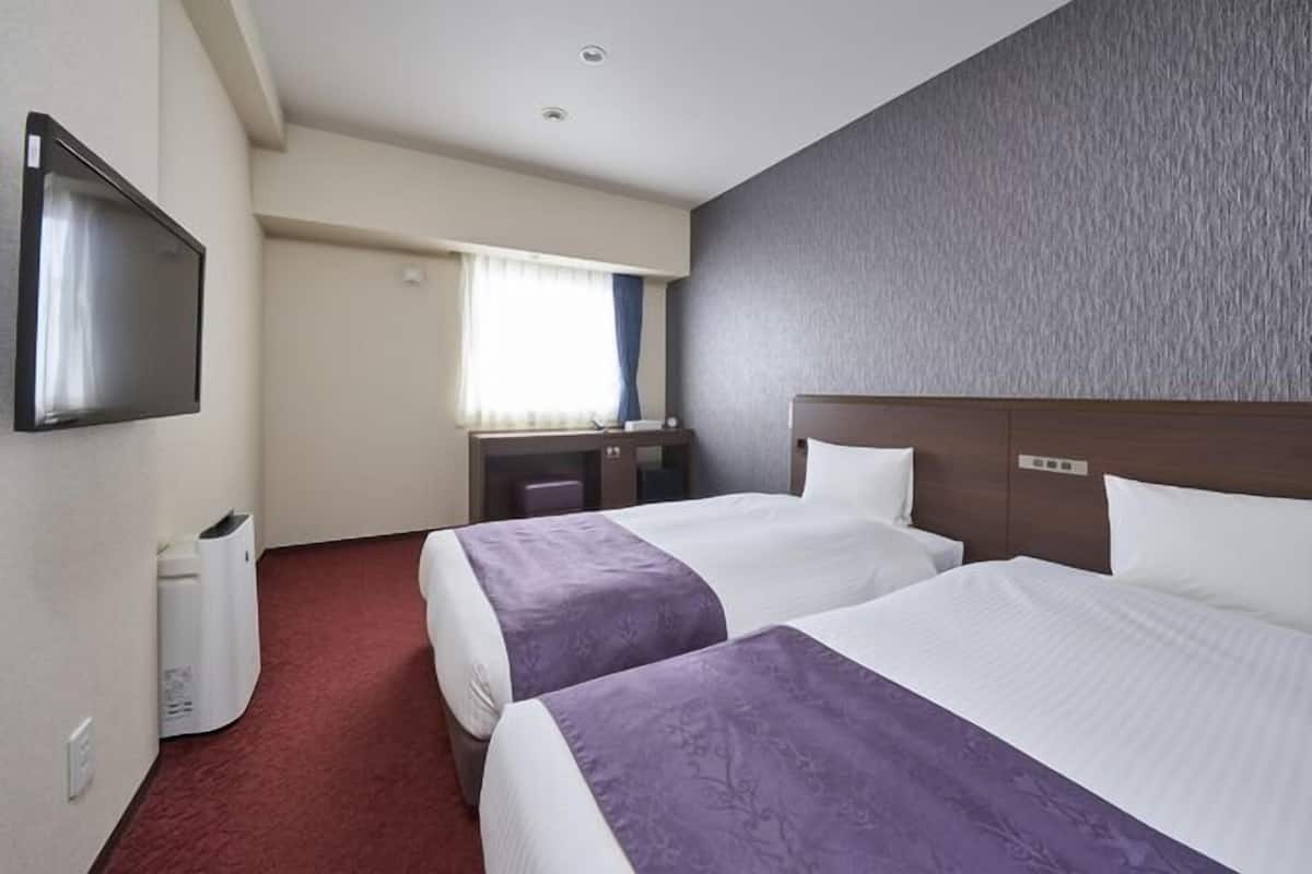 A hotel room with two beds, a TV mounted on the wall, a desk, and a window with curtains.