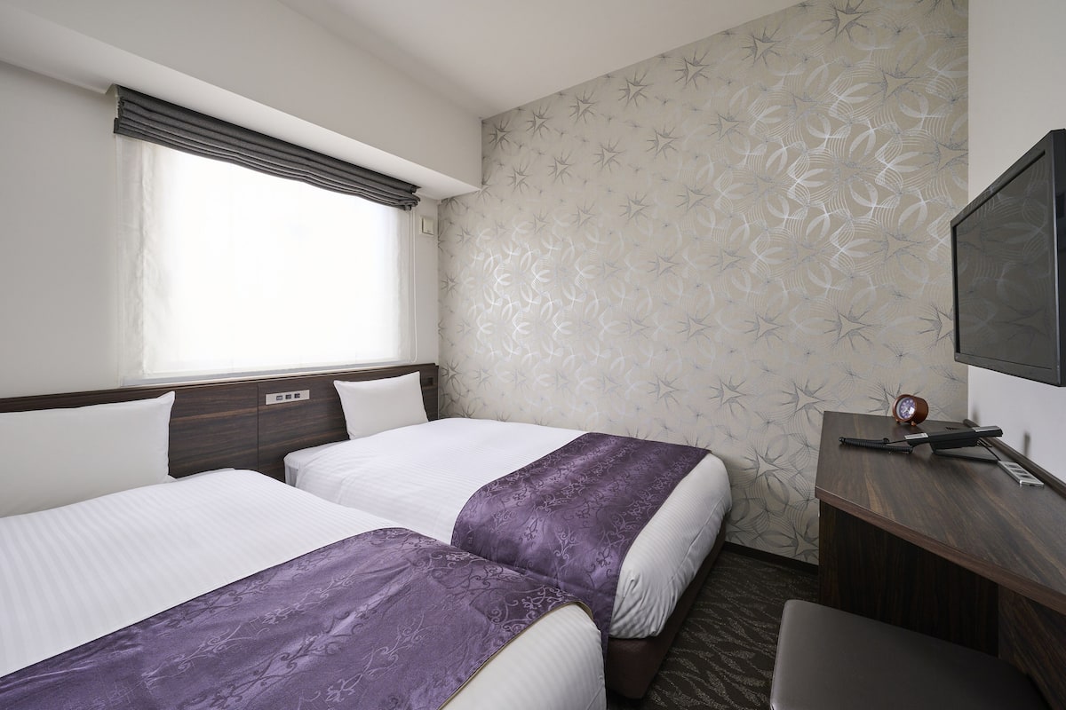 A hotel room with two beds, a desk, a television, and a window with curtains.