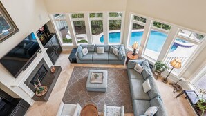 A spacious living room with a gray sectional sofa, a flat-screen TV, and a fireplace. Large windows offer views of a swimming pool and outdoor greenery. - Beautiful & Spacious 5br Home w/ Private Pool (Hilton Head Island)