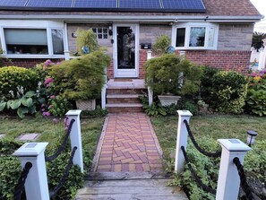 Ein Haus mit Ziegelaußenwand, einer Haustür, einer kleinen Veranda und einem Garten mit verschiedenen Pflanzen und Blumen.