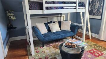 Un letto a castello con un divano blu sottostante, un tavolino rotondo con libri e un telecomando, e un tappeto con motivi floreali sul pavimento.