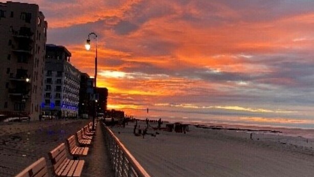 Un muelle con bancos a lo largo de una playa, edificios al fondo y un cielo al atardecer.