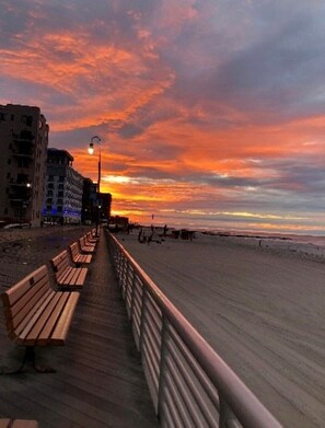 Eine Promenade mit Bänken entlang eines Strandes, im Hintergrund Gebäude und ein Sonnenuntergangshimmel.