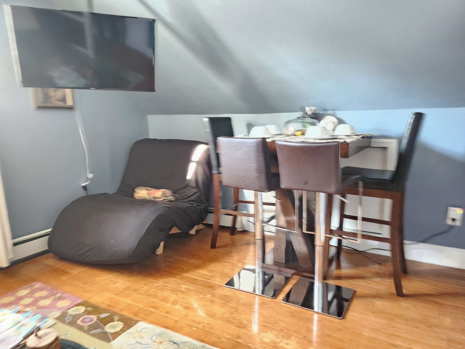 A room with a dining table, two chairs, a black sofa, a TV mounted on the wall, and a patterned rug on the floor.