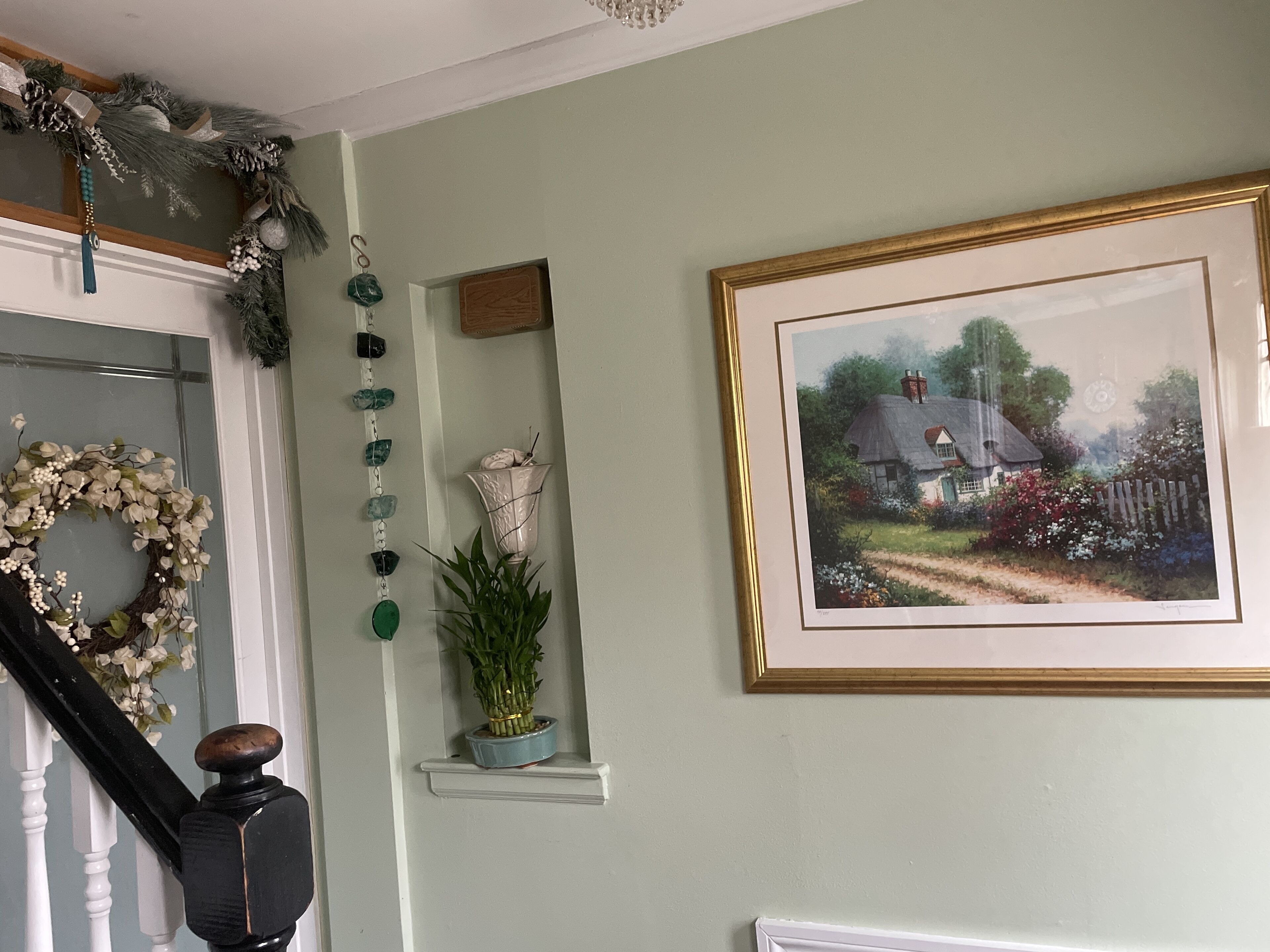 Un escalier avec une rampe noire et des balustrades blanches, un mur orné d’un tableau encadré représentant une maison de campagne, et une guirlande sur le côté gauche.