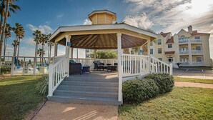 A covered outdoor area with steps leading to a seating area, surrounded by palm trees and a building with multiple balconies. - 2Bed2Bath, sleeps 6. Pet friendly. Pool. Hot tub. walking distance to the beach. (Corpus Christi)