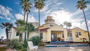 A yellow building with a porch, palm trees, and a bench. - 2Bed2Bath, sleeps 6. Pet friendly. Pool. Hot tub. walking distance to the beach. (Corpus Christi)