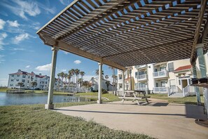 A covered outdoor area with a pool, palm trees, and multi-story buildings in the background. - 2Bed2Bath, sleeps 6. Pet friendly. Pool. Hot tub. walking distance to the beach. (Corpus Christi)