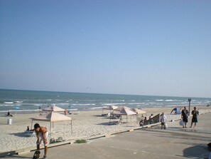 A beach with umbrellas, a concrete walkway, and the ocean in the background. - 2Bed2Bath, sleeps 6. Pet friendly. Pool. Hot tub. walking distance to the beach. (Corpus Christi)