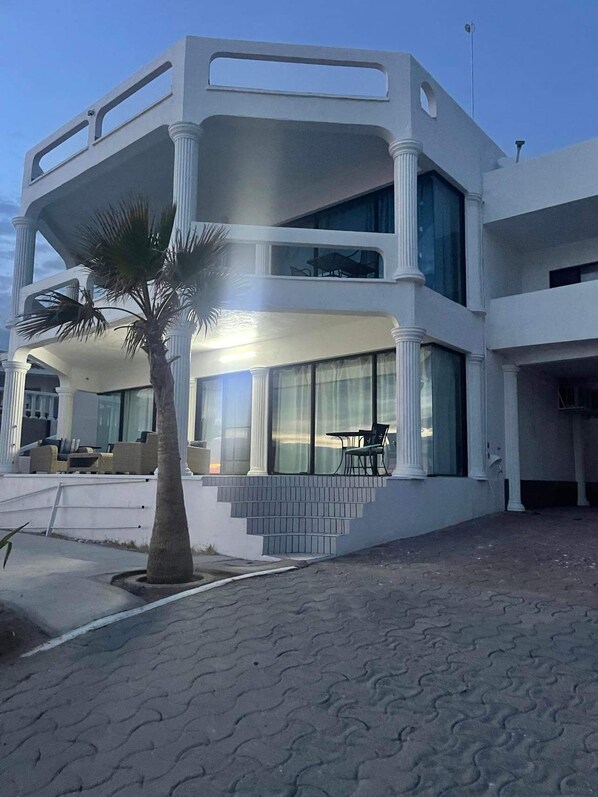 A modern two-story building with a symmetrical facade, large windows, and a palm tree in the foreground. - Oceanfront 10 bdrm 5 kitchen. Rent 6 bdrm Main House @ base price or add casitas (Puerto Peñasco)