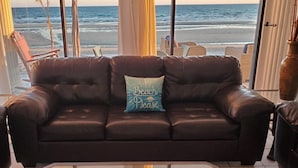 Ein dunkelbraunes Ledersofa mit Kissen, auf dem "Beach Please" steht, vor einem großen Fenster mit Blick auf einen Strand.