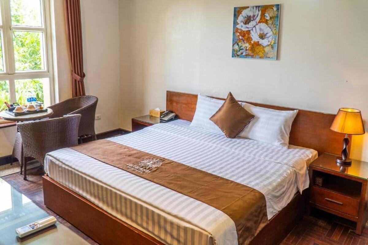 A hotel room with a large bed, a bedside table with a lamp, a chair, a small table with a tray of items, and a painting on the wall.