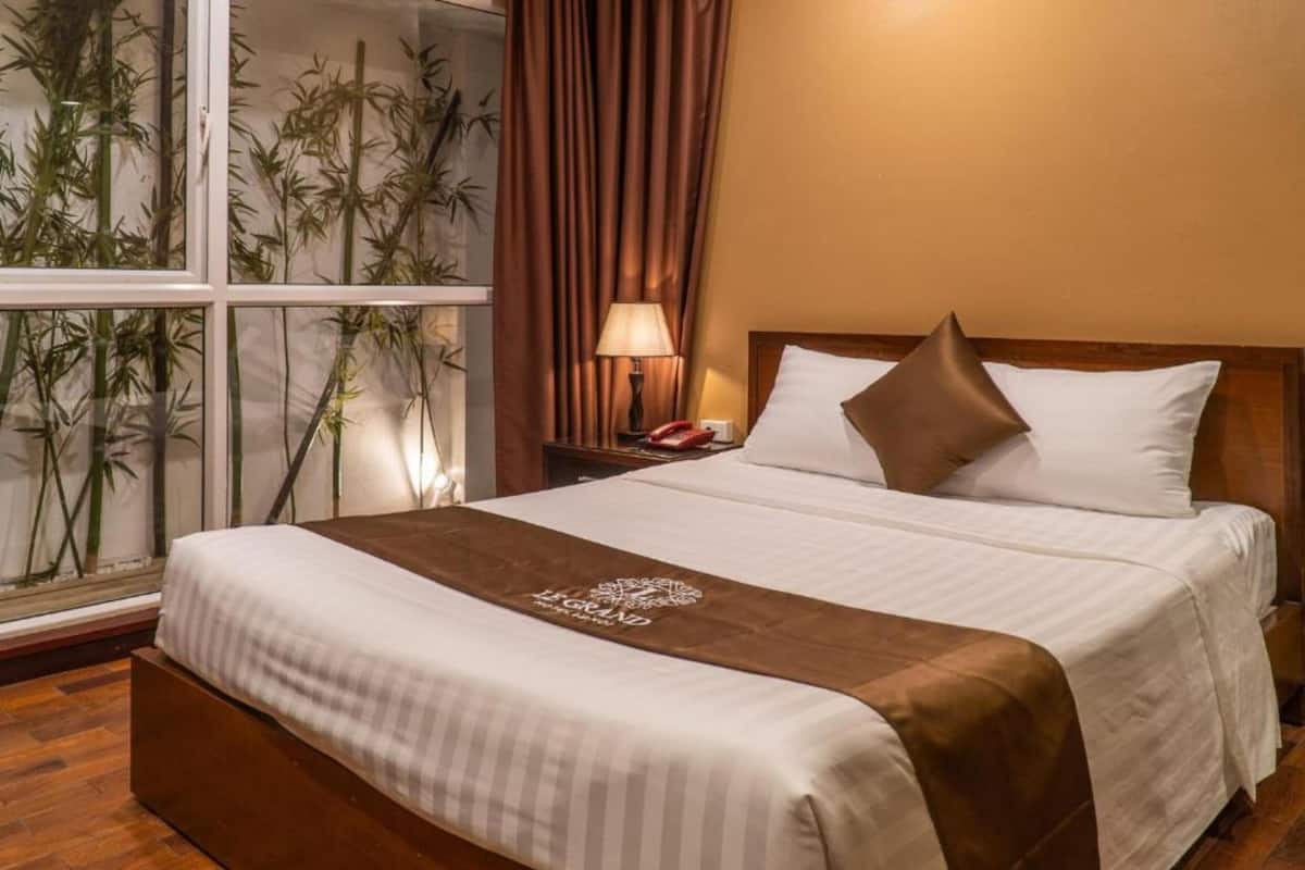 A hotel room with a bed, a nightstand, a lamp, and a window with bamboo plants.