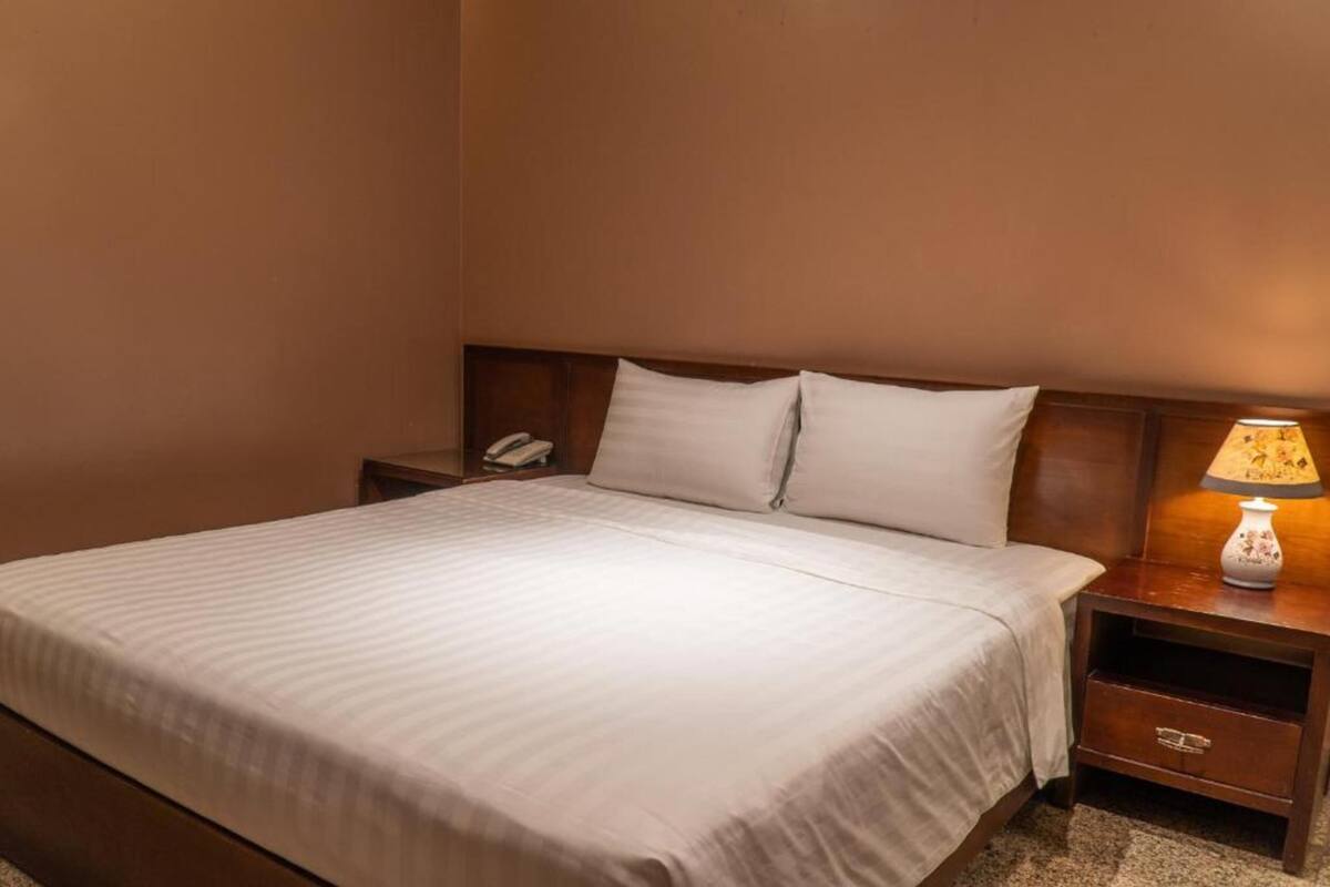 A neatly made bed with white linens, a wooden headboard, a bedside table with a lamp, and a phone.