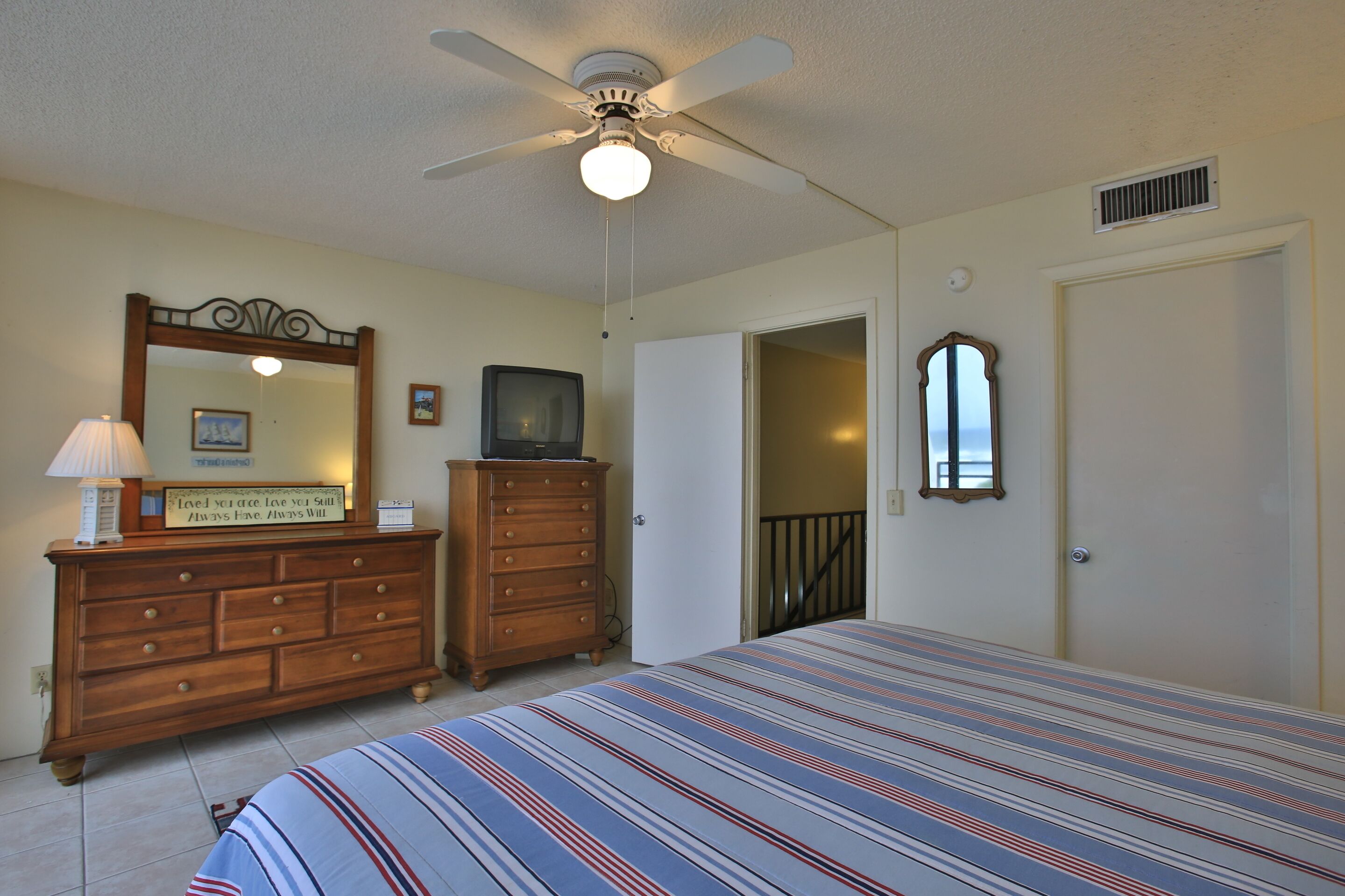 Une chambre à coucher comprenant un lit, une commode, un ventilateur de plafond et une porte donnant sur une autre pièce.