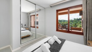 Quarto de hotel moderno com cama grande, porta de correr de vidro para uma varanda e vista para o verde lá fora.