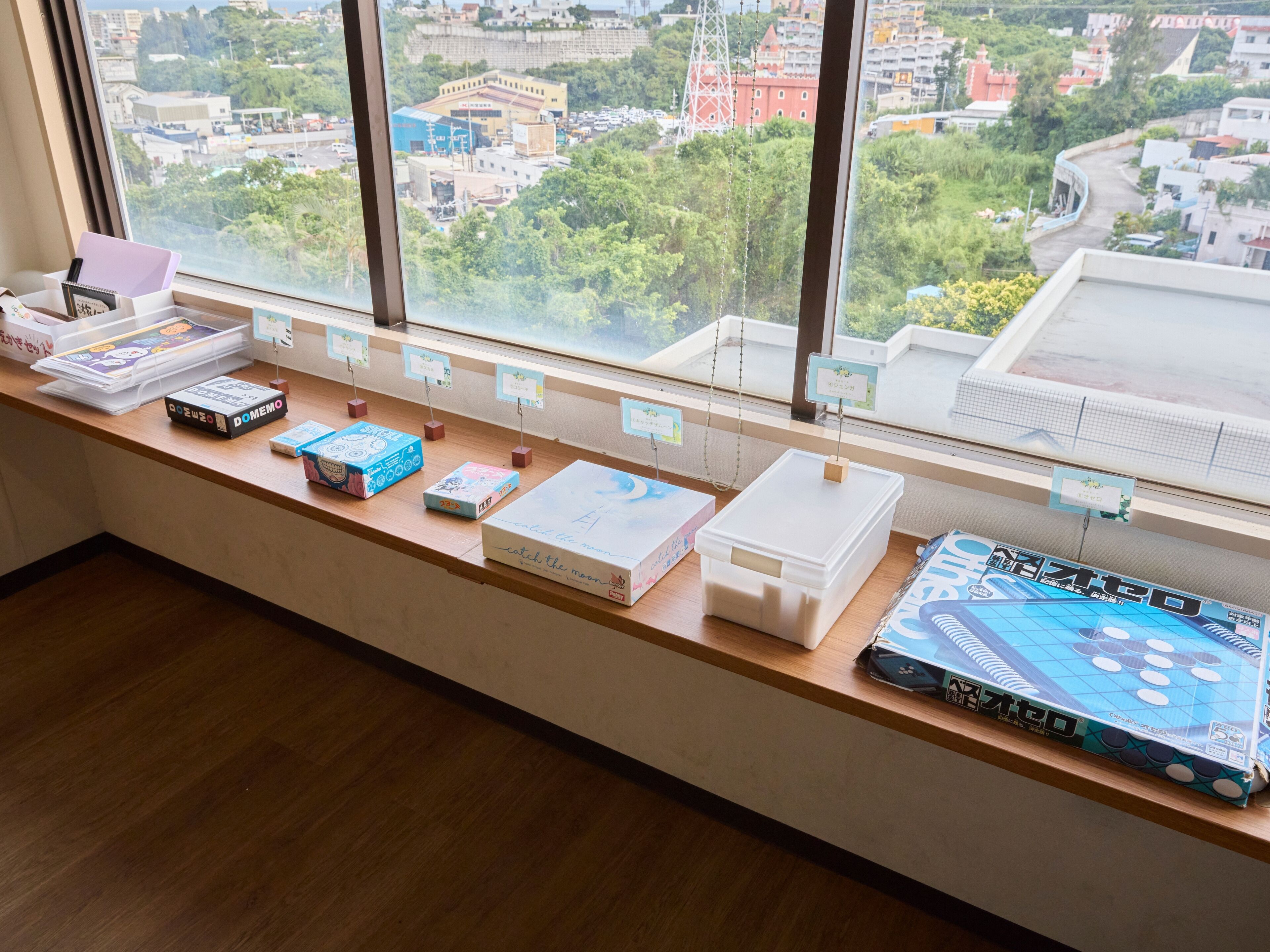 A long wooden table with various board games and boxes, set against a window with a city view.