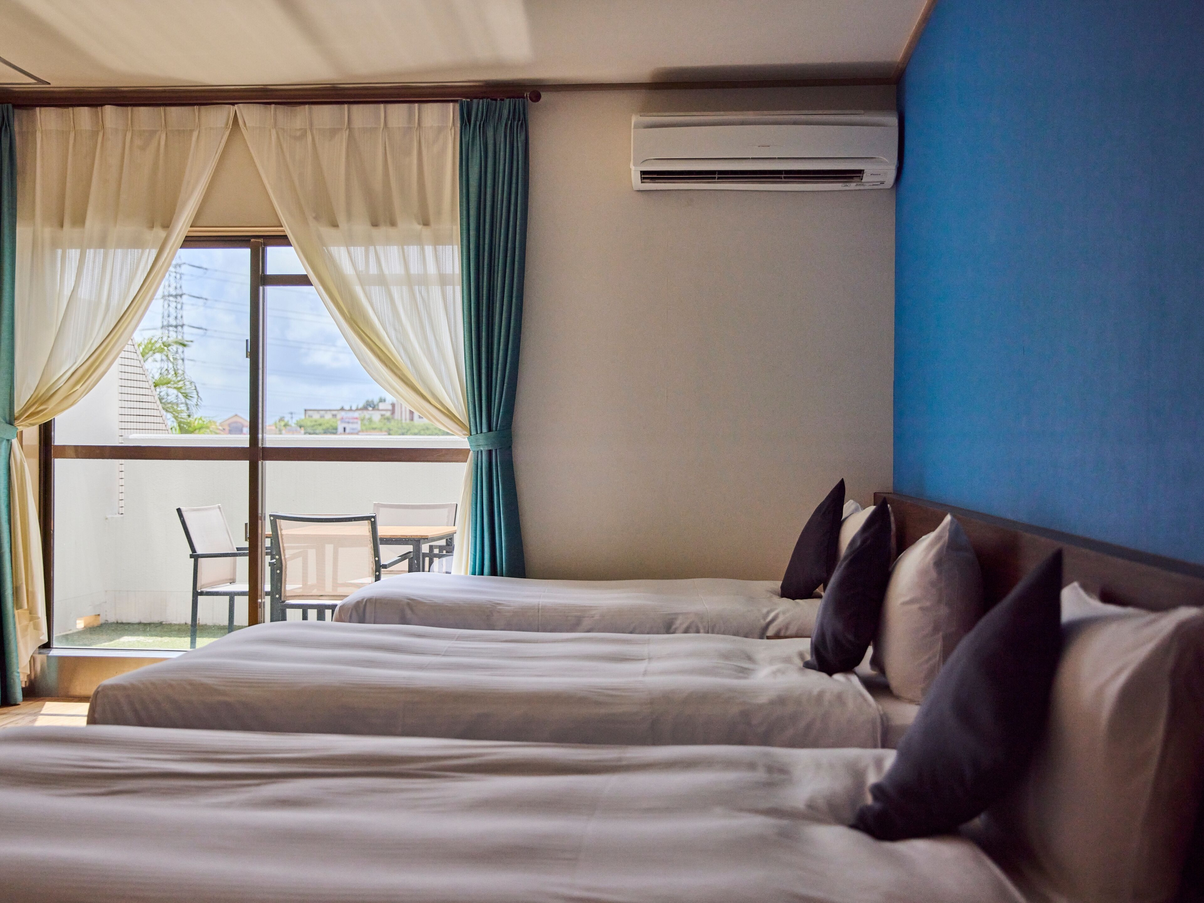 Habitación de hotel con dos camas, un balcón con mesa y sillas, y un equipo de aire acondicionado en la pared.