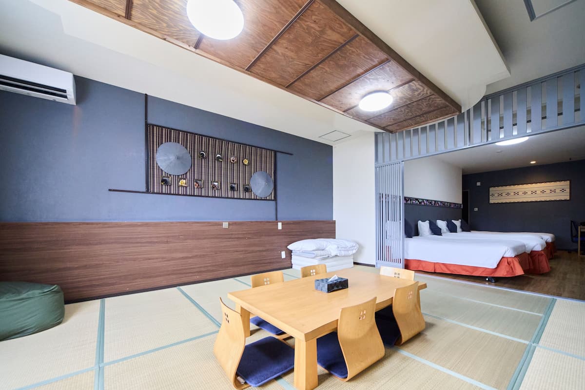 A hotel room with a tatami mat floor, a wooden table with chairs, a bed with white bedding, a wall-mounted air conditioner, and a ceiling with wooden panels and recessed lighting.