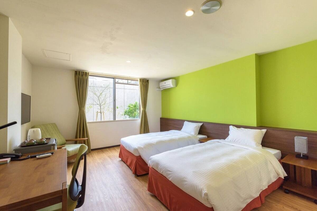 A hotel room with two beds, a desk with a chair, a window with curtains, and an air conditioning unit.
