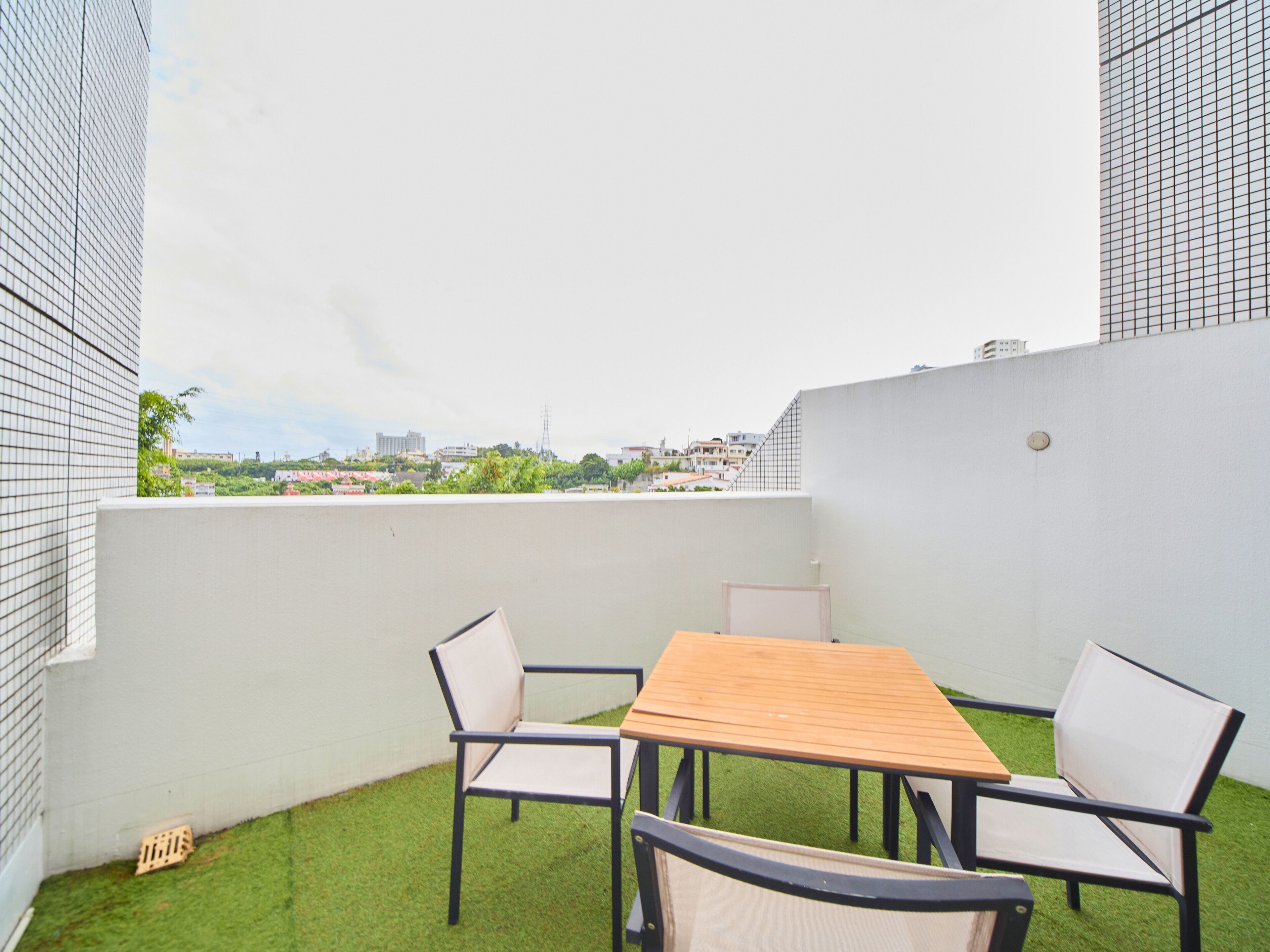 A rooftop terrace with a wooden table and chairs, surrounded by white walls and a view of the cityscape.