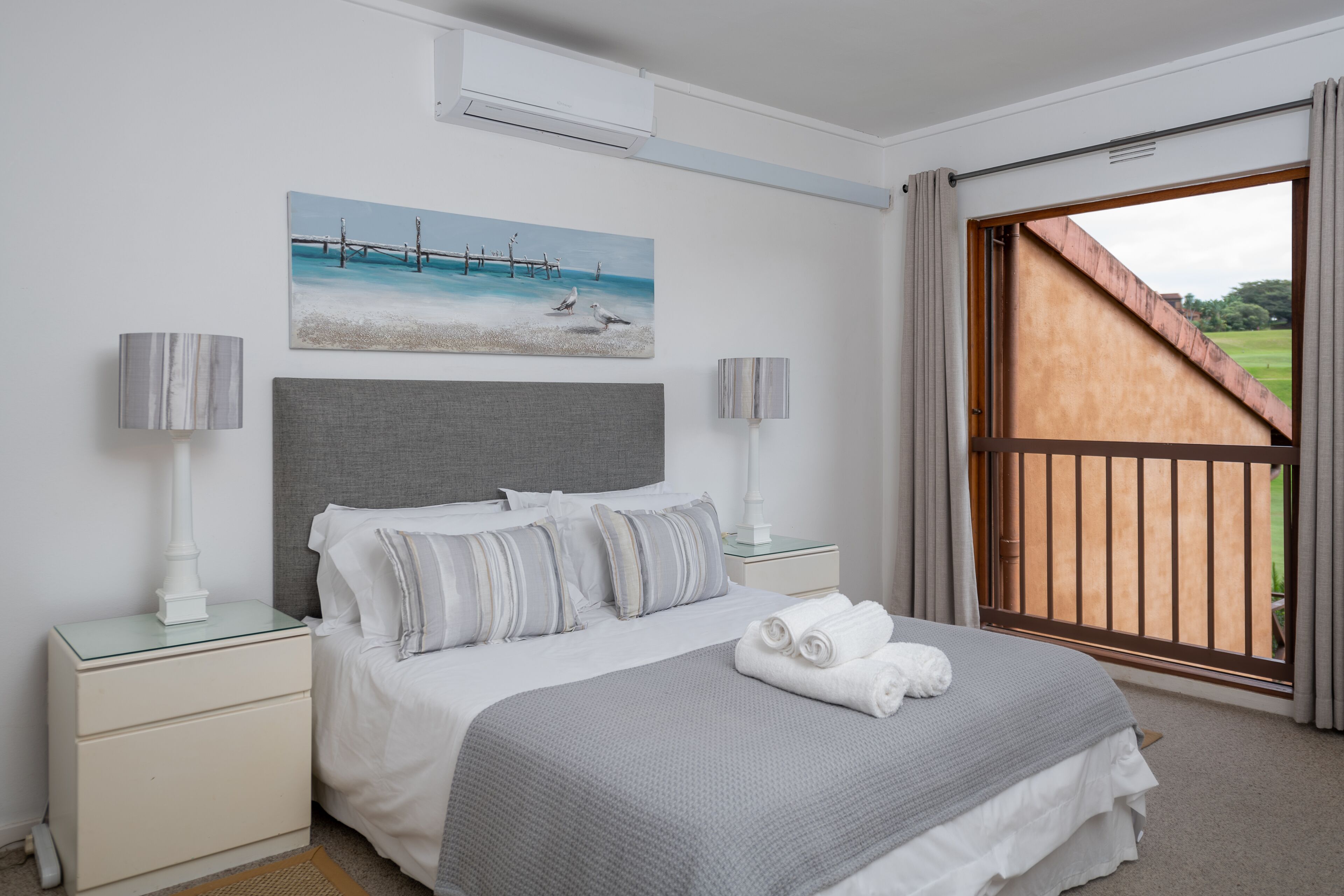 Une chambre à coucher avec un lit, des tables de chevet, un tableau et une vue sur l’extérieur, grâce à une grande fenêtre.